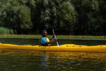 Person wearing a blue life jacket paddling a bright yellow kayak on calm water with a forested shoreline in the background