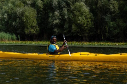 Person wearing a blue life jacket paddling a bright yellow kayak on calm water with a forested shoreline in the background