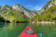 The tip of a red kayak on the smooth, green waters of Albania's Komani Lake with a backdrop of forest-finged rocky mountains and another red kayak in the distance.