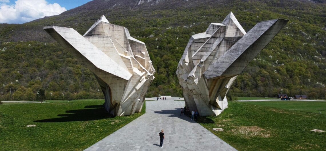 Massive concrete angular sculptures in a grassy park with a mountain backdrop; people walk along a paved path, foregrounded by a wide walkway.