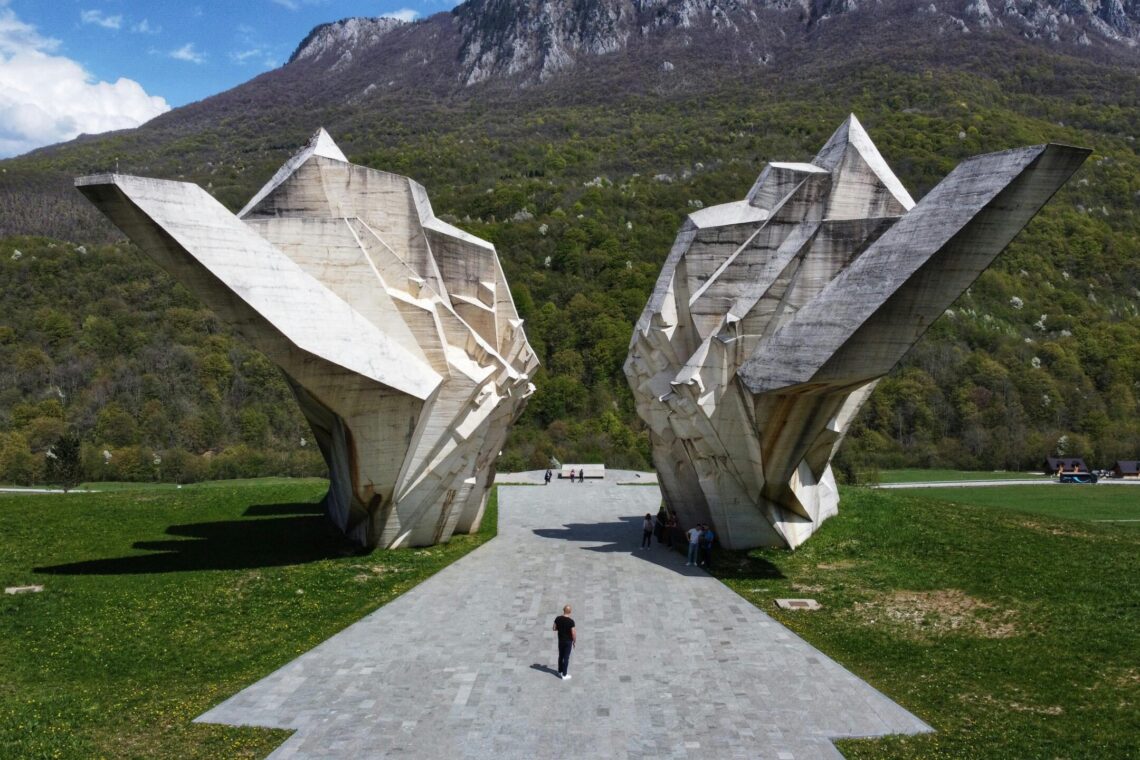 Massive concrete angular sculptures in a grassy park with a mountain backdrop; people walk along a paved path, foregrounded by a wide walkway.