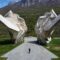 Massive concrete angular sculptures in a grassy park with a mountain backdrop; people walk along a paved path, foregrounded by a wide walkway.
