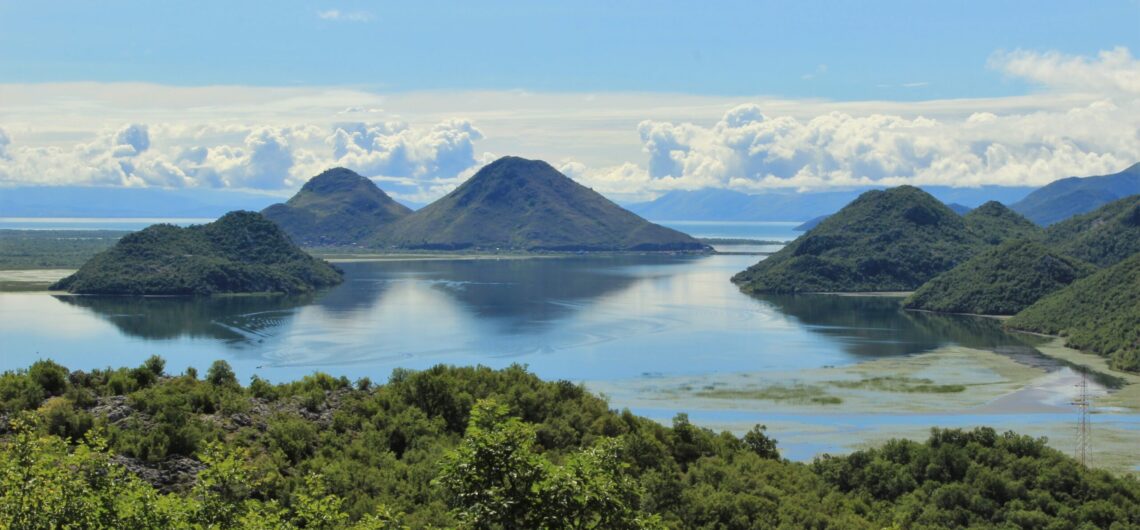 Wide coastal landscape with green hills rising from calm blue water and scattered clouds in a bright sky, distant mountains on the horizon.