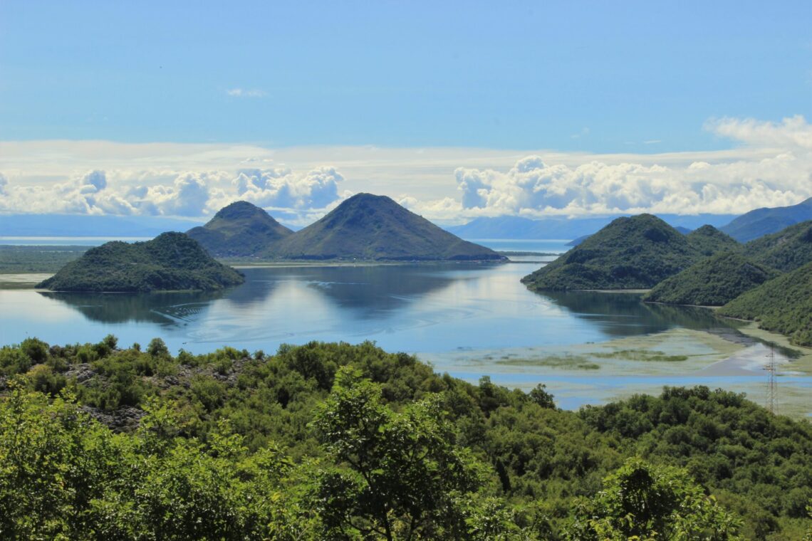 Wide coastal landscape with green hills rising from calm blue water and scattered clouds in a bright sky, distant mountains on the horizon.