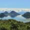Wide coastal landscape with green hills rising from calm blue water and scattered clouds in a bright sky, distant mountains on the horizon.