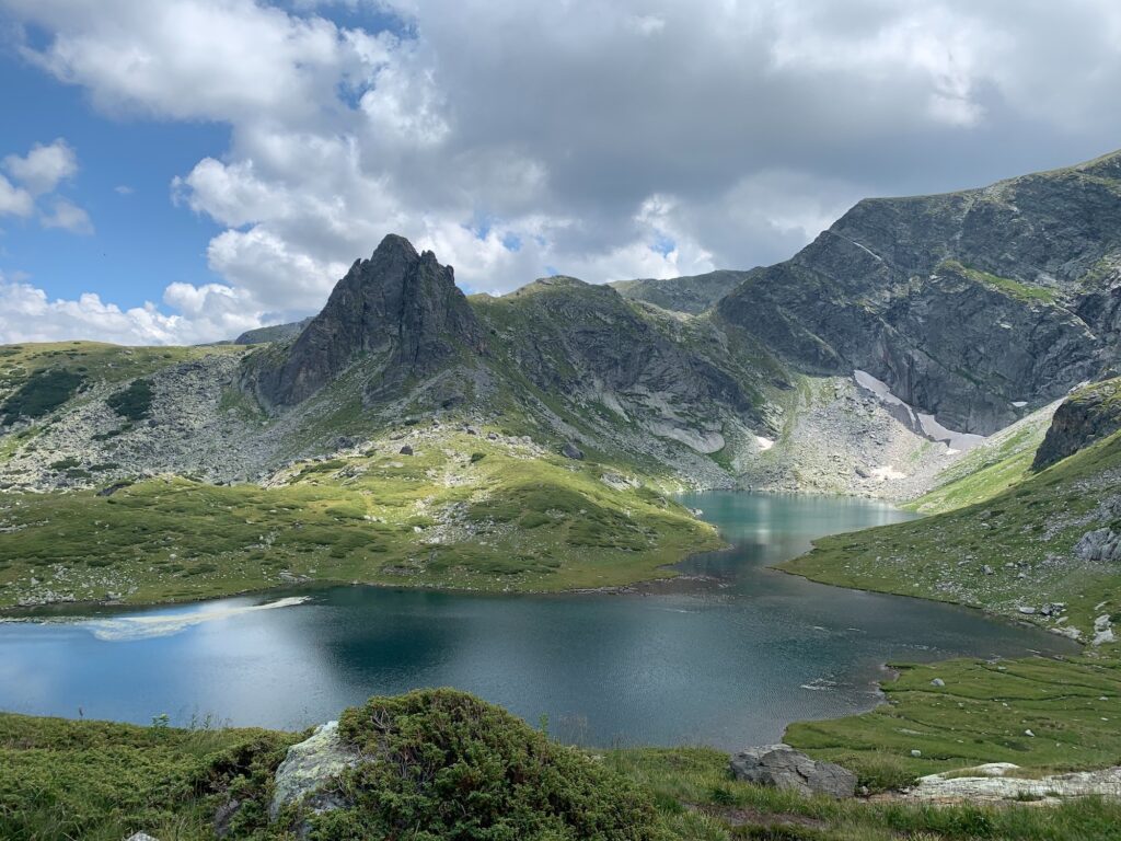 Bulgaria mountain landscape with a jagged rocky peak, green slopes, and a blue-green lake under a partly cloudy sky. seven rila lakes hiking circuit.
