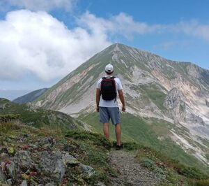 Hiker with backpack standing on a rocky mountain trail, facing a green-sloped peak under a blue sky.