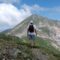 Hiker with backpack standing on a rocky mountain trail, facing a green-sloped peak under a blue sky.