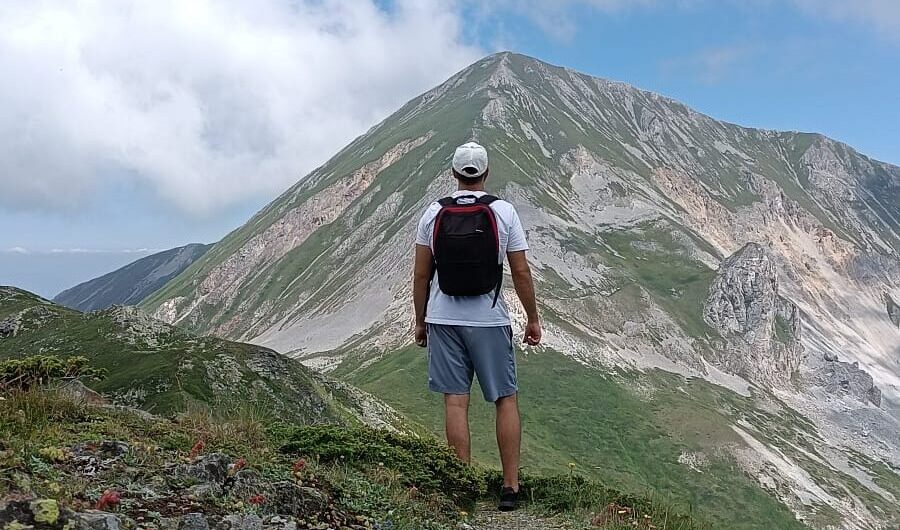 Hiker with backpack standing on a rocky mountain trail, facing a green-sloped peak under a blue sky.