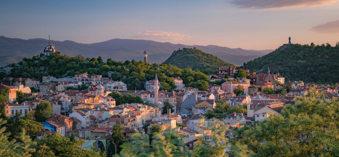 Sunlit hillside town with red roofs, green trees, and distant mountains at sunset.