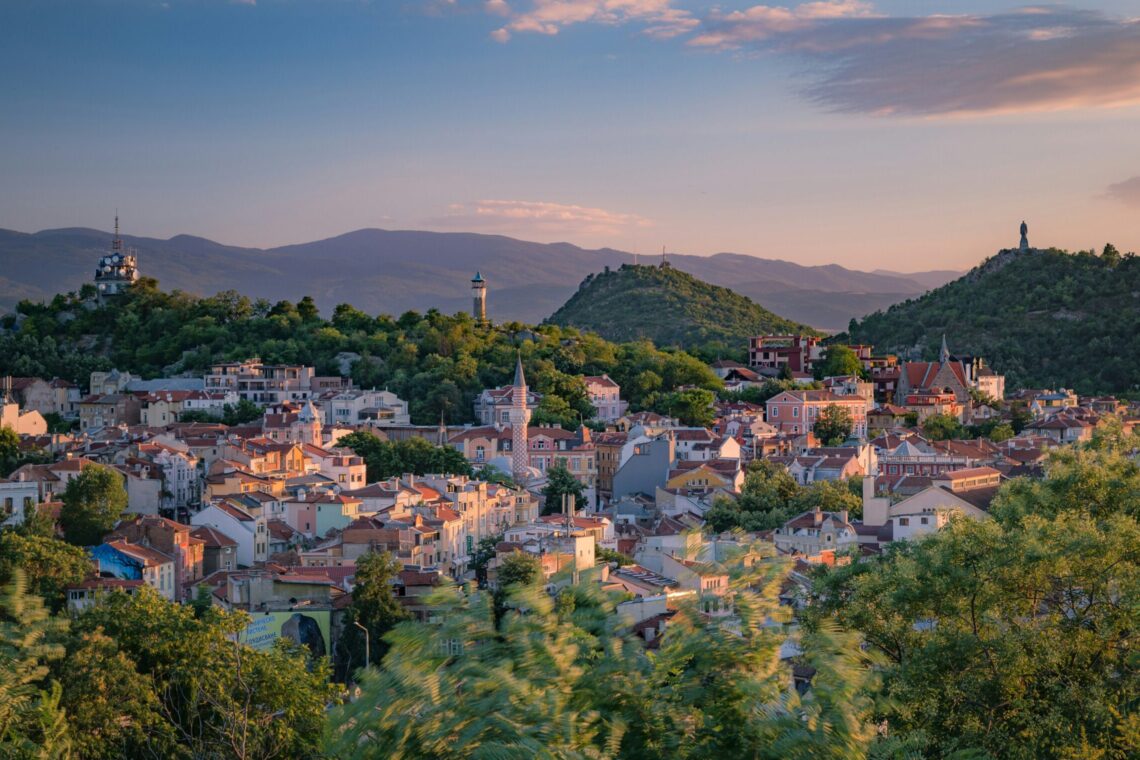 Sunlit hillside town with red roofs, green trees, and distant mountains at sunset.