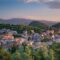 Sunlit hillside town with red roofs, green trees, and distant mountains at sunset.