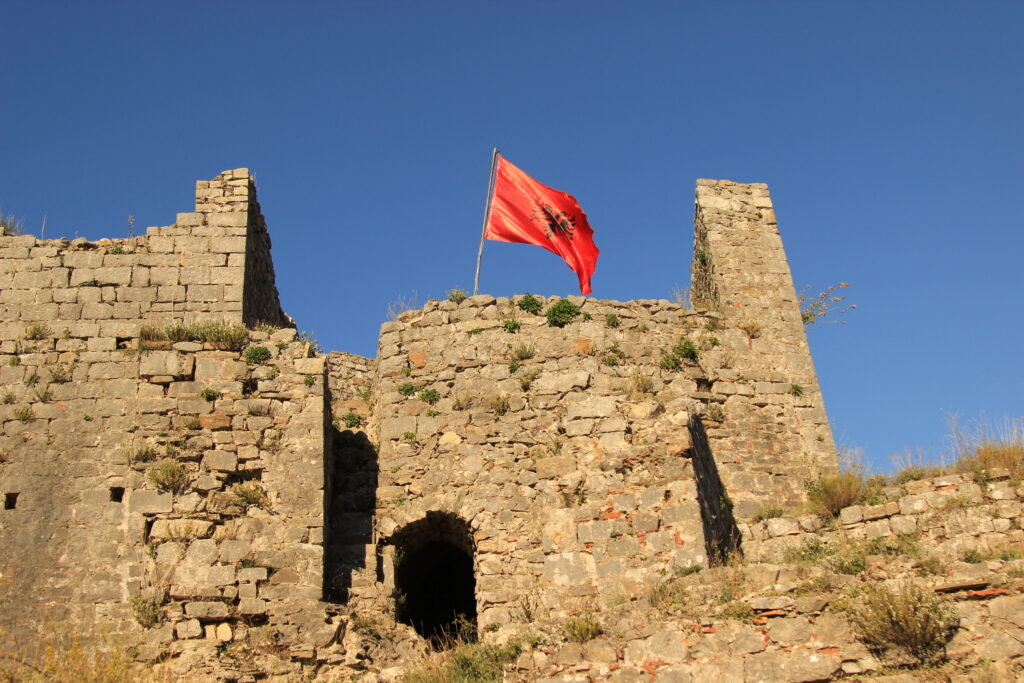 Red Albanian flag flying on a blue-sky day above ancient stone castle walls with grass and weathered stones.