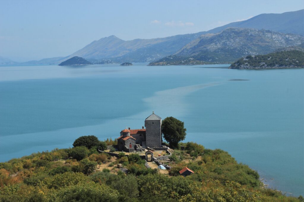 A view onto a medieval island monastery in the foreground, with Lake Shkodra and mountains behind it.