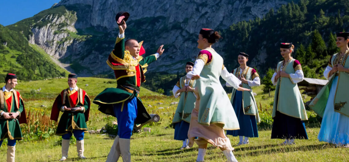 Group of people in colorful traditional folk costumes dancing in a grassy meadow with mountains in the background.