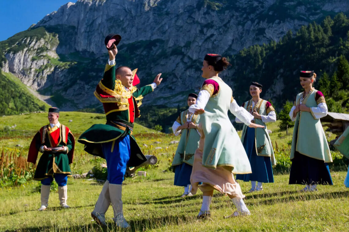 Group of people in colorful traditional folk costumes dancing in a grassy meadow with mountains in the background.