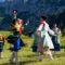 Group of people in colorful traditional folk costumes dancing in a grassy meadow with mountains in the background.