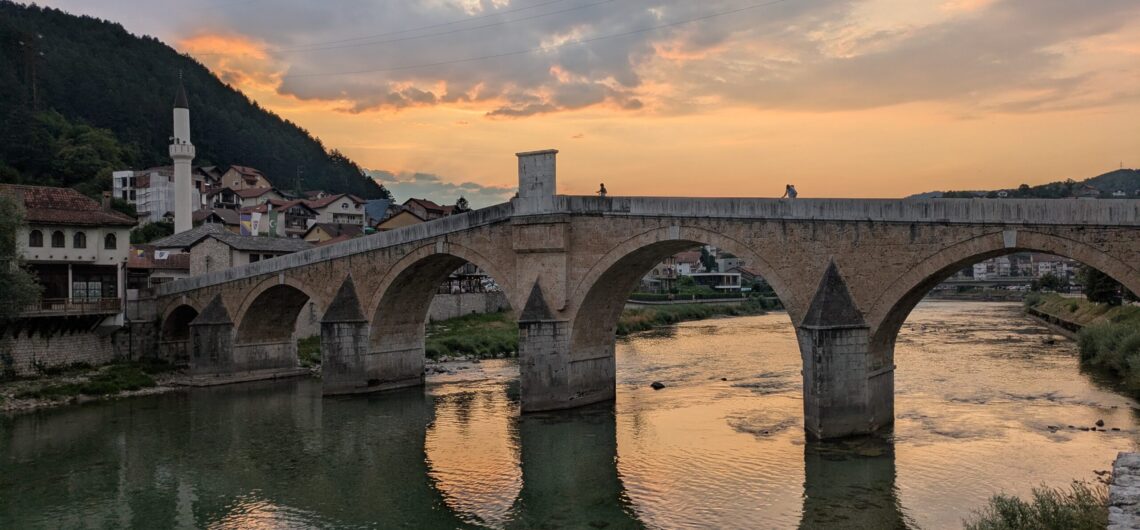 Stone arched bridge over a calm river at sunset, with a hillside village and warm orange sky in the background, two people standing on the bridge.