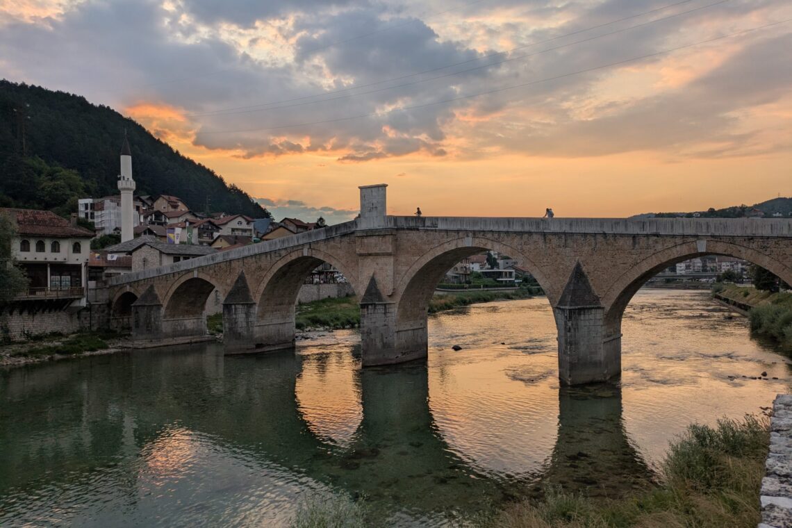 Stone arched bridge over a calm river at sunset, with a hillside village and warm orange sky in the background, two people standing on the bridge.