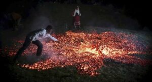 Man in a white shirt pushes glowing embers on the ground with a long stick at night, while a person in red traditional clothing watches in the background.