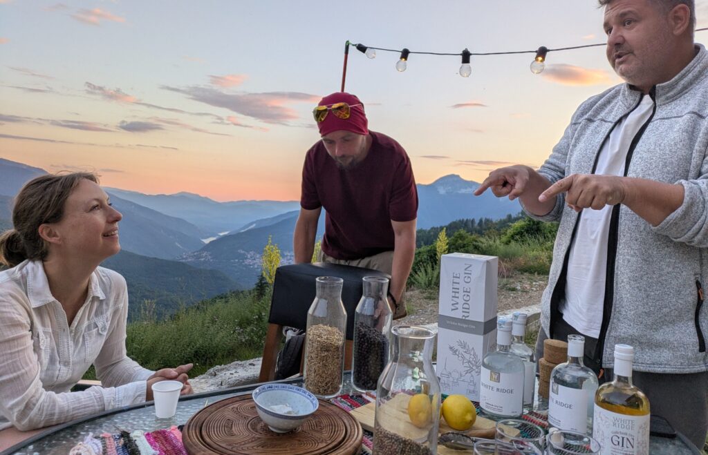 Group of people around a table with gin bottles and lemons outdoors at sunset, overlooking mountains