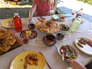Outdoor meal table with braided bread centerpiece, bowls of olives and salads, yellow patterned plates, drink jugs, and a person in a pink shirt reaching for food.