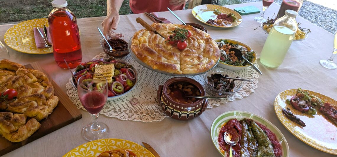 Outdoor meal table with braided bread centerpiece, bowls of olives and salads, yellow patterned plates, drink jugs, and a person in a pink shirt reaching for food.