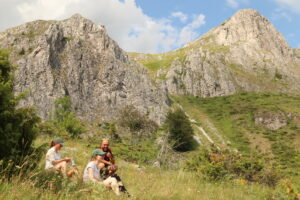 Group of hikers taking a break in a grassy valley with rugged gray rock cliffs in the background under a blue sky.