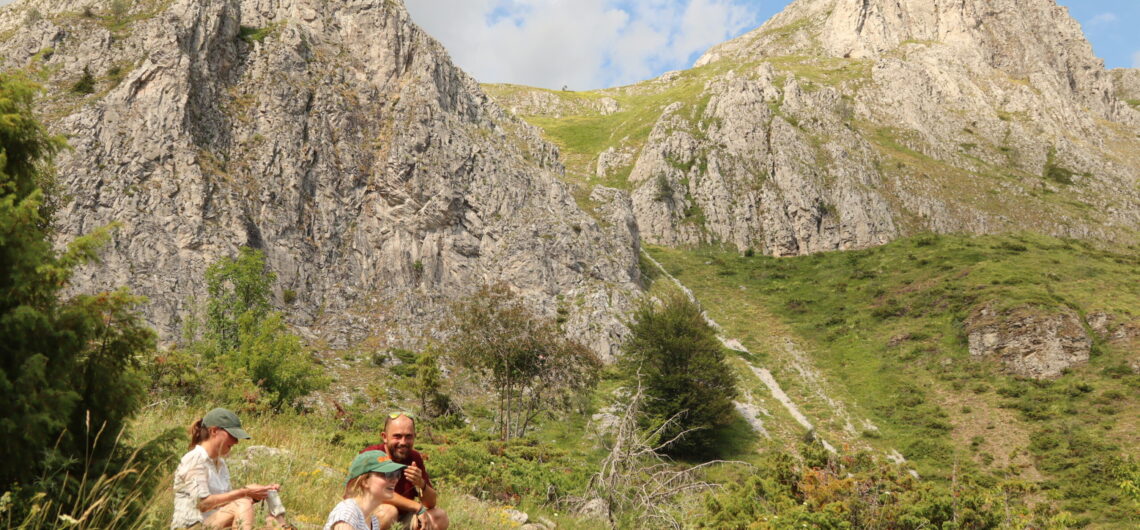 Group of hikers taking a break in a grassy valley with rugged gray rock cliffs in the background under a blue sky.